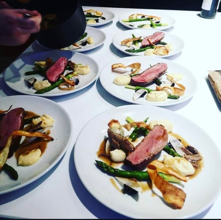 Multiple plates of plated duck or steak with roasted vegetables and potatoes on a white table during a fine dining service