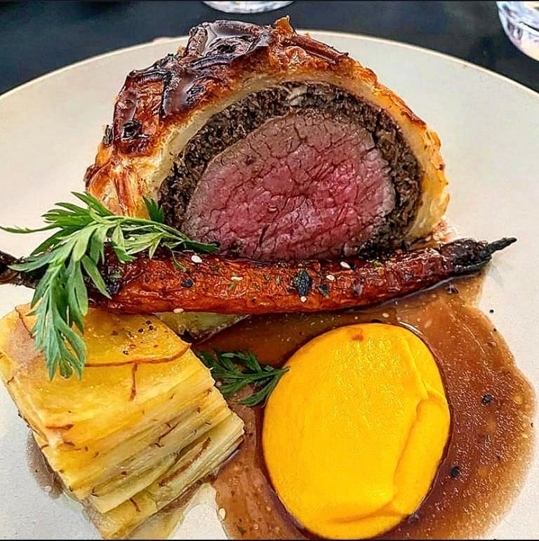Sliced beef Wellington with rare pink center, served with roasted carrot, layered potato cake, and herbs on a white plate